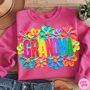 May include: A pink sweatshirt with the word "GRANDMA" in colorful, raised letters, surrounded by vibrant, textured flowers in shades of blue, yellow, orange, and pink. The sweatshirt is made of a soft material.