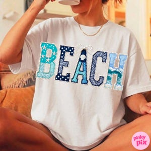 May include: White t-shirt with the word "BEACH" in a decorative, patchwork style. The letters are in shades of blue and turquoise, with pearl-like embellishments. The "B" features a star design.