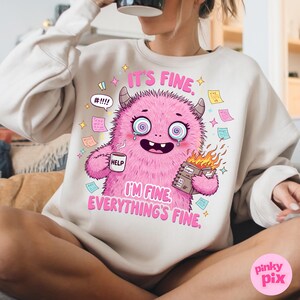 May include: A white sweatshirt with a pink monster graphic. The monster is holding a coffee mug that says "Help" and a book on fire. The text on the sweatshirt says "It's fine. I'm fine. Everything's fine."