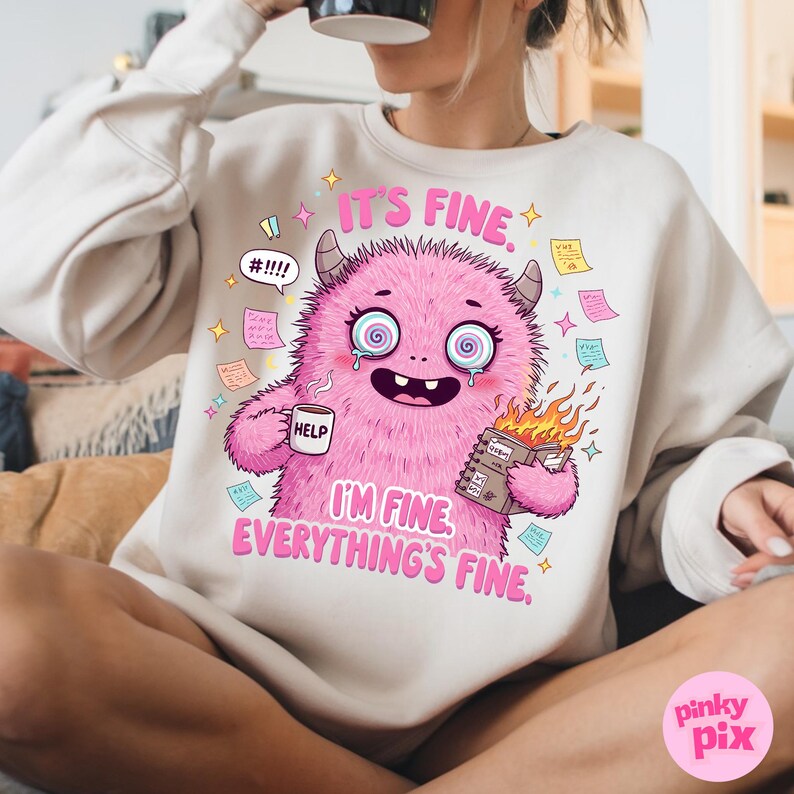 May include: A white sweatshirt with a pink monster graphic. The monster is holding a coffee mug that says "Help" and a book on fire. The text on the sweatshirt says "It's fine. I'm fine. Everything's fine."