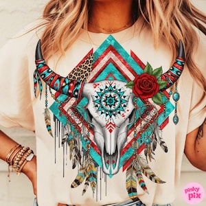 Op de afbeelding: Crèmekleurig T-shirt met een gedetailleerde afbeelding van een stierschedel. Het ontwerp bevat turquoise, rode en witte geometrische patronen, een rode roos, dromenvangers en veren, wat een bohemien esthetiek creëert.