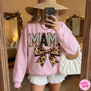 May include: A light pink sweatshirt with the word "MAM" in jeweled letters and a leopard print bow. The sweatshirt is long-sleeved and has a crew neck. The image also shows white shorts and a straw hat.