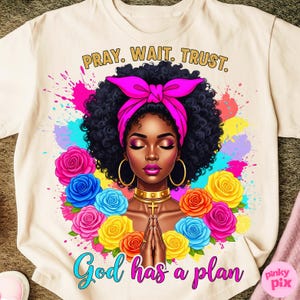 May include: Cream-colored t-shirt featuring a vibrant illustration of a Black woman with closed eyes, a pink headscarf, and gold hoop earrings. The design includes colorful roses, paint splatters, and the text "PRAY. WAIT. TRUST." and "God has a plan."