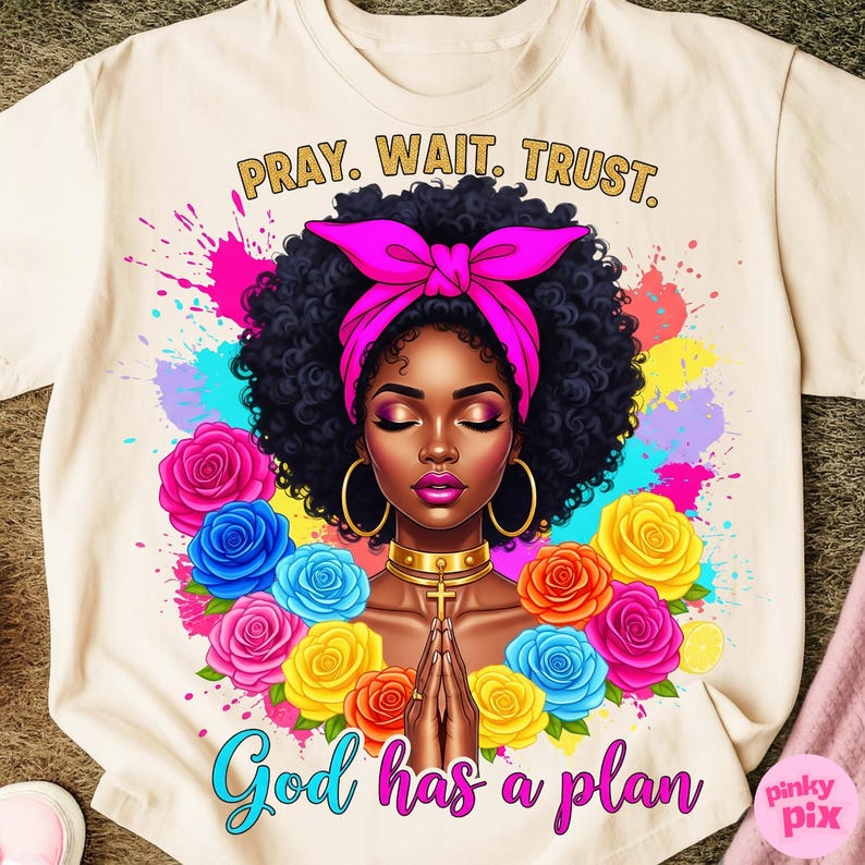 May include: Cream-colored t-shirt featuring a vibrant illustration of a Black woman with closed eyes, a pink headscarf, and gold hoop earrings. The design includes colorful roses, paint splatters, and the text "PRAY. WAIT. TRUST." and "God has a plan."