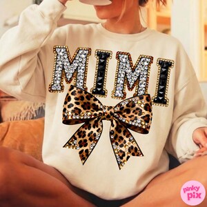 May include: Cream-colored sweatshirt with the word "MIMI" in jeweled letters and a leopard print bow. The bow is embellished with rhinestones. The sweatshirt is a casual, comfortable style.