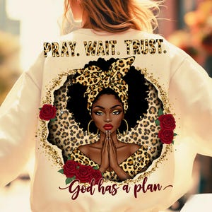 May include: A cream-colored sweatshirt with a black and gold leopard print design. The design features a woman with a leopard print headband, praying with her hands clasped together. The text "Pray. Wait. Trust. God has a plan" is printed on the sweatshirt.