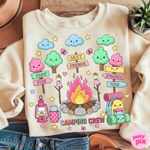 May include: Cream-colored sweatshirt with a camping-themed graphic. The design includes a campfire, trees, clouds, and the words "Camping Crew," "Best Friends," "Camp Fun," and "S'mores Time." The graphic is in a cartoon style with pastel colors.