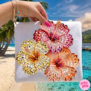 May include: White square pillow featuring three jeweled hibiscus flowers in red, yellow, and orange. The flowers are embellished with sparkling crystals. The background shows a tropical beach scene with palm trees and turquoise water.