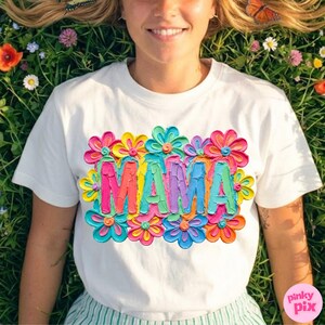 May include: White t-shirt with a colorful floral design and the word "MAMA" in vibrant, textured lettering. The design features a variety of bright colors, including pink, yellow, blue, and green, creating a cheerful and eye-catching look.