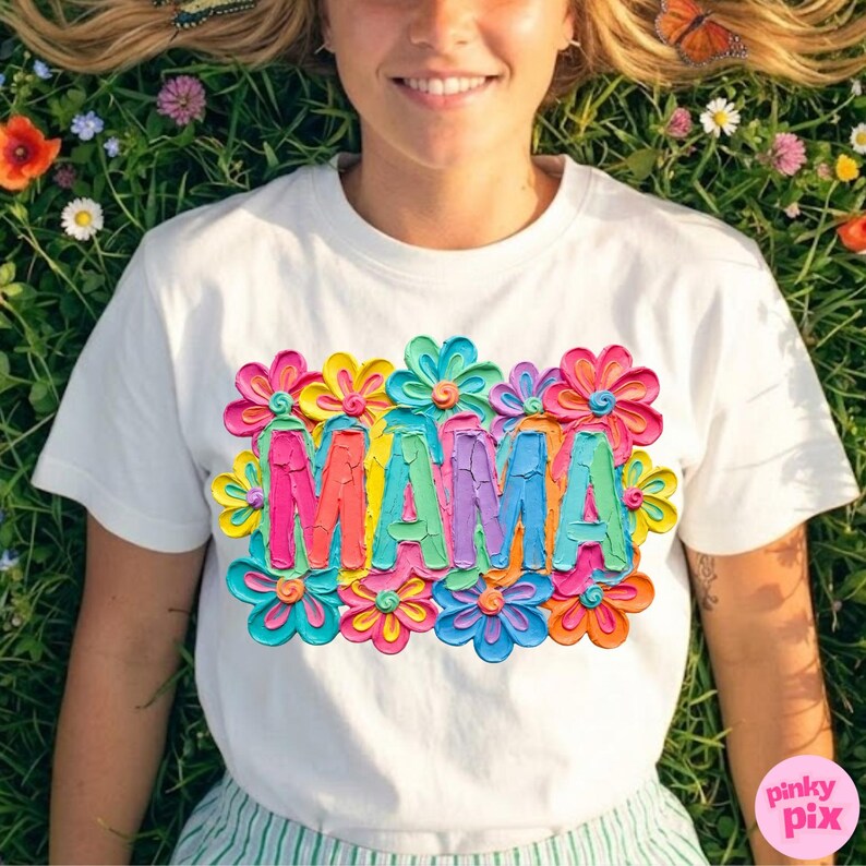 May include: White t-shirt with a colorful floral design and the word "MAMA" in vibrant, textured lettering. The design features a variety of bright colors, including pink, yellow, blue, and green, creating a cheerful and eye-catching look.
