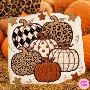 May include: A white sweatshirt featuring five embroidered pumpkins with varied patterns including black and white harlequin, leopard print, brown polka dots, and plaid, accented with small brown stars and swirling vine details.