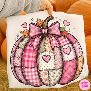 May include: A white pumpkin-shaped design with a pink bow, hearts, and autumn leaves. The pumpkin is made of various pink patterned patches, including gingham, floral, and polka dots. The design is held up by a hand.