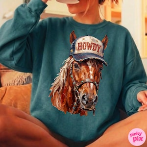 May include: Teal-colored sweatshirt featuring a detailed illustration of a horse wearing a 'HOWDY' cap. The horse has a brown coat, a white mane, and a bridle with turquoise accents. The cap is navy blue with white lettering and gold stars.