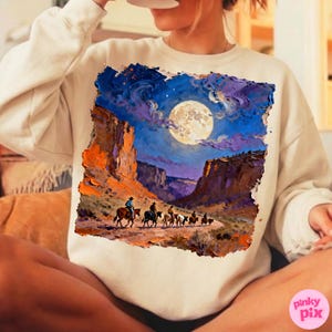 May include: Cream-colored sweatshirt featuring a vibrant painting of a desert landscape under a full moon. The artwork depicts cowboys on horseback riding through a canyon, with rich blues, oranges, and purples dominating the scene.