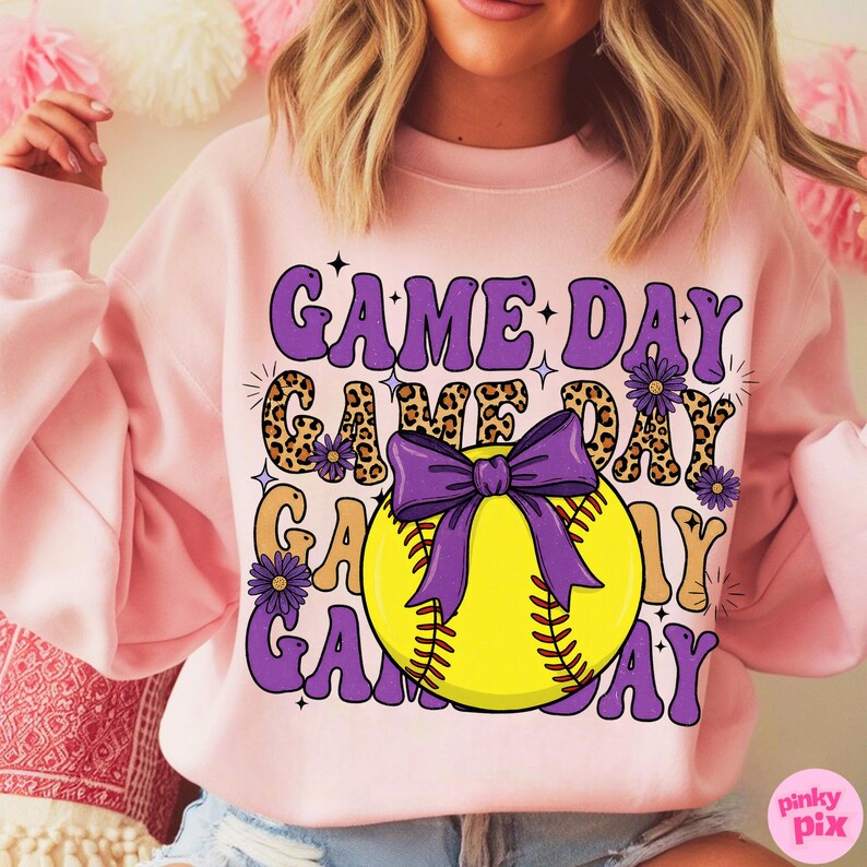 May include: Pink sweatshirt with the words "GAME DAY" in purple, leopard print, and yellow. A yellow softball with a purple bow is in the center. The sweatshirt has long sleeves.