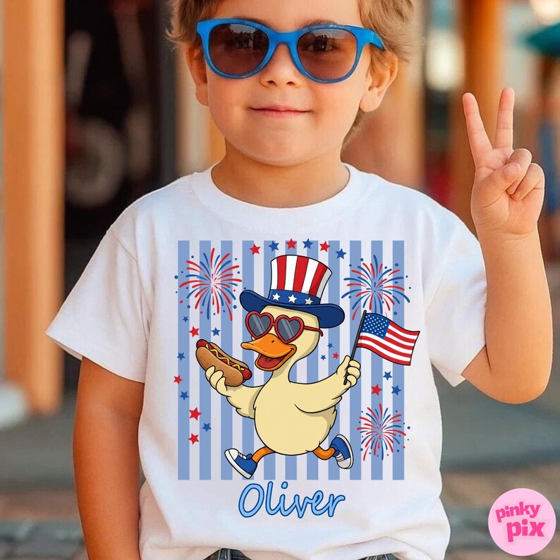 May include: White t-shirt with a patriotic duck graphic. The duck wears heart-shaped sunglasses, a top hat, and holds a hot dog and an American flag. Blue stripes and fireworks surround the duck. The name "Oliver" is written in blue.