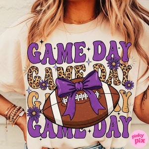 May include: Cream-colored t-shirt with a football-themed graphic. The design features the words "GAME DAY" in purple, a brown football with a purple bow, and leopard print accents. The shirt is a casual, sporty style.