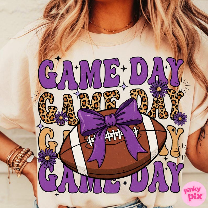May include: Cream-colored t-shirt with a football-themed graphic. The design features the words "GAME DAY" in purple, a brown football with a purple bow, and leopard print accents. The shirt is a casual, sporty style.