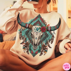 May include: A cream sweatshirt with a detailed bull skull graphic. The design incorporates turquoise, red, and brown elements, a red rose, and feathers. The graphic is set against a geometric background. The horns are long and curved.