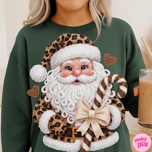 May include: Green sweatshirt featuring a whimsical Santa Claus design. Santa is depicted with a white beard, rosy cheeks, and a leopard print hat and coat. He holds a candy cane and a cream-colored bow. Two heart-shaped cookies are also visible.