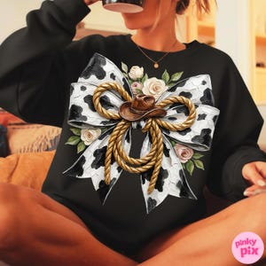 May include: Black sweatshirt featuring a large bow design with a cow print pattern, a brown cowboy hat, and rope detailing. The bow is adorned with white and pink roses and green leaves. The sweatshirt is a comfortable, casual style.