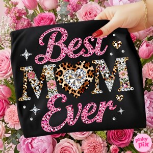 May include: Black t-shirt with the text "Best MOM Ever" in pink, leopard print, and rhinestone lettering. The design includes a leopard print heart with a large crystal in the center, surrounded by pink flowers and diamonds. The shirt is held in front of a background of pink roses.