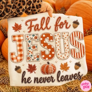May include: A white sweatshirt featuring the embroidered text Fall for JESUS he never leaves. The word JESUS is composed of large letters with varied patterns including orange gingham, floral, polka dots, and stripes, accented by leaves and acorns.