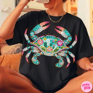 May include: Black t-shirt featuring a detailed, colorful crab design. The crab is adorned with turquoise, pink, and white jewel-like accents, and floral patterns. The design is centered on the shirt.