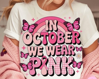 Könnte beinhalten: Weißes T-Shirt mit dem Text "IN OCTOBER WE WEAR PINK" in rosa und schwarzer Schrift. Das Design umfasst einen rosa Regenbogen, Schmetterlinge und ein rosa Band. Das Shirt ist ein lässiger Stil.