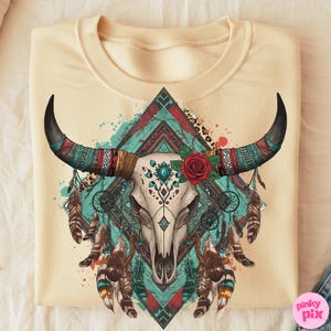 May include: Cream-coloured sweatshirt with a detailed graphic of a bull skull adorned with turquoise, red, and brown tribal patterns. The design includes a red rose, feathers, and dream catchers, creating a bohemian aesthetic.