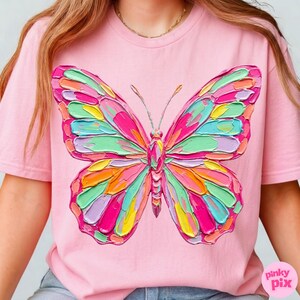 May include: Pink t-shirt featuring a colorful butterfly design. The butterfly has a textured, painted appearance with shades of pink, green, yellow, and orange. The shirt has short sleeves and a crew neck. The text "pinky pix" is in the bottom right corner.