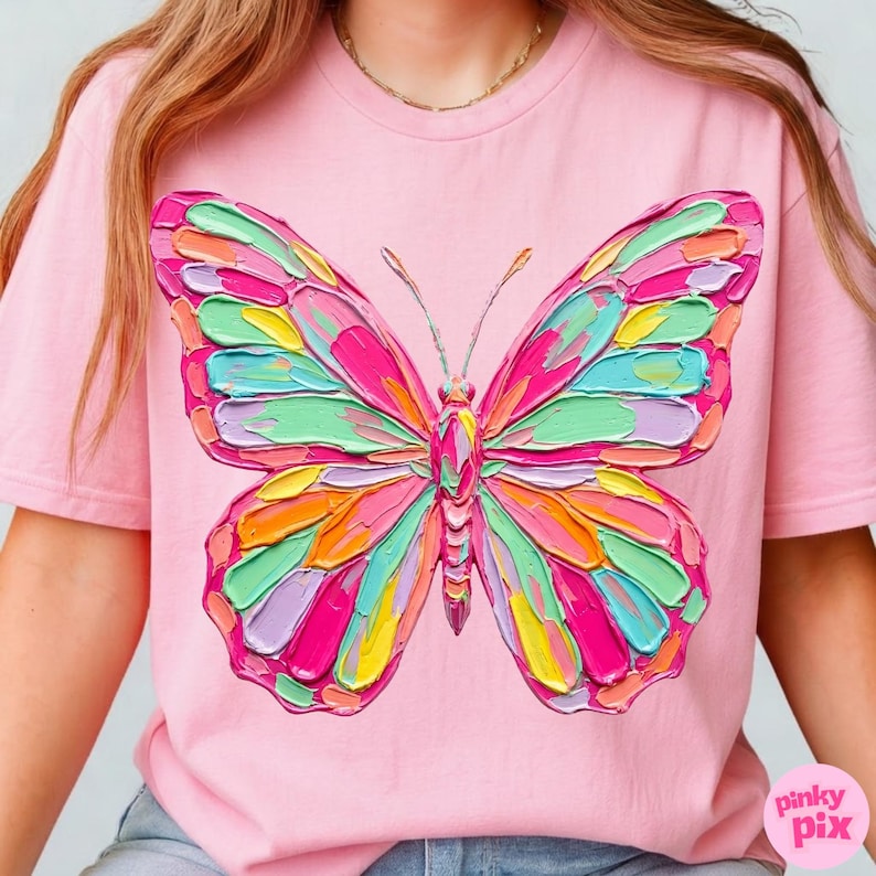 May include: Pink t-shirt featuring a colorful butterfly design. The butterfly has a textured, painted appearance with shades of pink, green, yellow, and orange. The shirt has short sleeves and a crew neck. The text "pinky pix" is in the bottom right corner.
