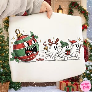 Pode incluir: Uma fronha branca com um design de desenho animado com tema natalino. Um enfeite zangado persegue três fantasmas, um com chifres de rena, um com um gorro verde e um com um gorro de Papai Noel, cada um carregando presentes ou bengalas doces. O enfeite é vermelho e verde.