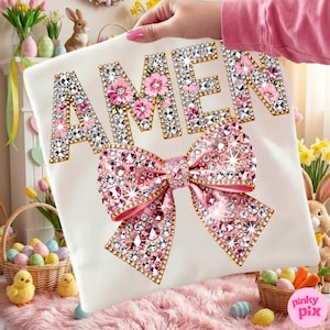 May include: White t-shirt featuring the word "AMEN" in sparkling rhinestones and pink floral details. A large pink rhinestone bow is positioned below the text. The shirt is held up by a person.