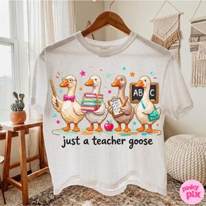 May include: White t-shirt featuring a whimsical illustration of four cartoon geese dressed as teachers. The geese are holding books, a clipboard, and a pointer. The text "just a teacher goose" is printed below the geese.