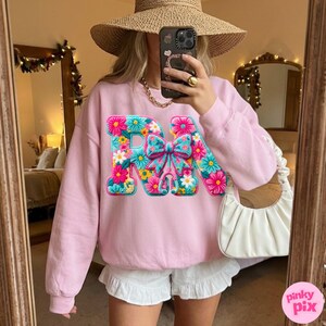May include: A light pink sweatshirt with the letters "RN" in a floral design. The letters are decorated with colorful flowers and a bow. The sweatshirt is long-sleeved and has a crew neck.