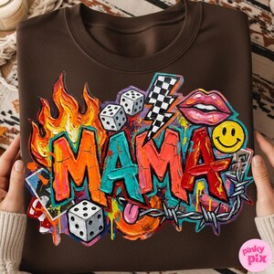 May include: Brown sweatshirt with a colorful graphic design. The design features the word "MAMA" in graffiti style, flames, dice, a lightning bolt, lips, a smiley face, and barbed wire.