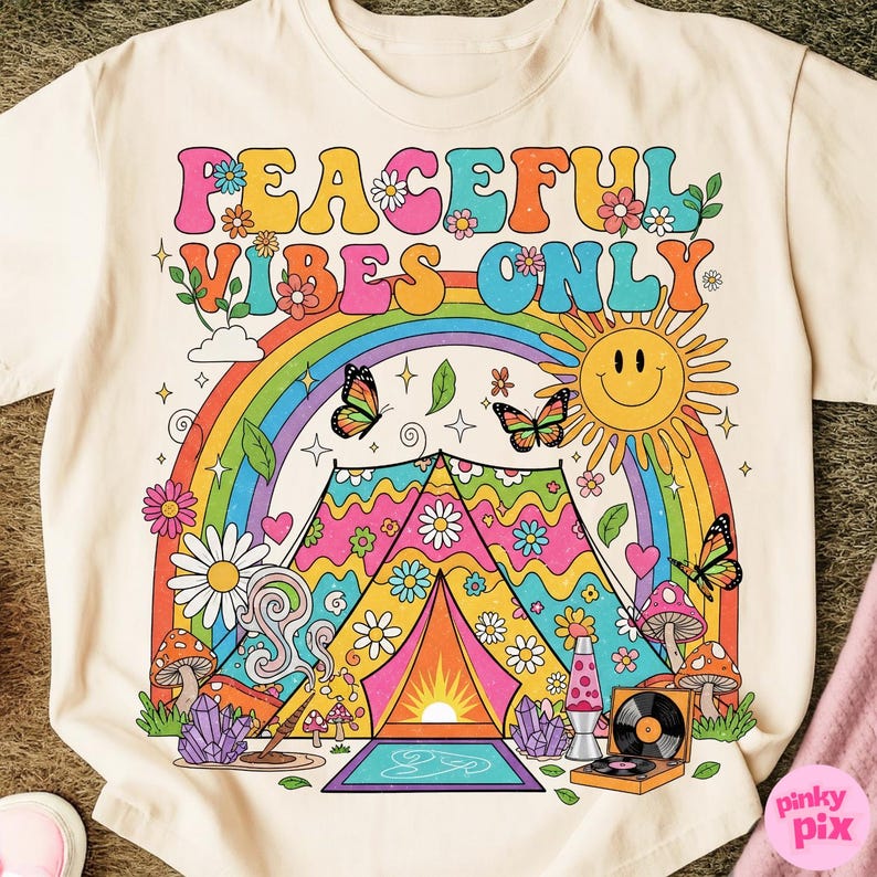 May include: Cream-colored t-shirt with a colorful graphic design. The design features the text "PEACEFUL VIBES ONLY" above a rainbow, butterflies, a smiling sun, and a tent. Additional elements include flowers, mushrooms, and a record player.