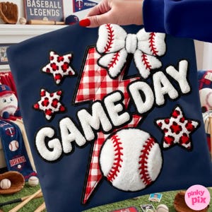 Pode incluir: Almofada azul marinho com tema de beisebol. Apresenta um laço branco, estrelas com estampa de leopardo vermelho, uma bola de beisebol e as palavras "GAME DAY" em branco. Um raio xadrez vermelho e branco também está presente.