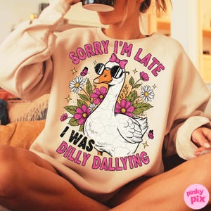 K&ouml;nnte beinhalten: Beiges Sweatshirt mit dem Text "SORRY I'M LATE I WAS DILLY DALLYING" in Pink. Die Grafik zeigt eine wei&szlig;e Gans mit Sonnenbrille und rosa Schleife, umgeben von Blumen und Schmetterlingen. Das Sweatshirt ist ein l&auml;ssiger Stil.