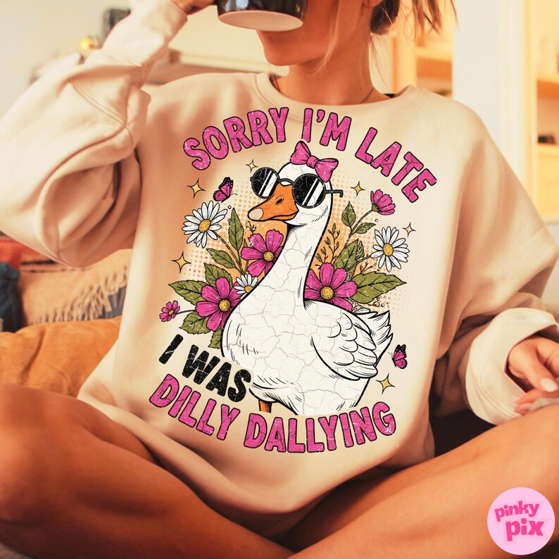 K&ouml;nnte beinhalten: Beiges Sweatshirt mit dem Text "SORRY I'M LATE I WAS DILLY DALLYING" in Pink. Die Grafik zeigt eine wei&szlig;e Gans mit Sonnenbrille und rosa Schleife, umgeben von Blumen und Schmetterlingen. Das Sweatshirt ist ein l&auml;ssiger Stil.