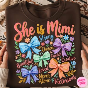 K&ouml;nnte beinhalten: Ein braunes Sweatshirt mit dem Text "She is Mimi" in Rosa und Gelb, umgeben von bunten Schleifen und floralen Akzenten. Die W&ouml;rter "Strong, Chosen, Capable, Creative, Beautiful, Loved, Never Alone, Victorious" sind ebenfalls zu sehen.