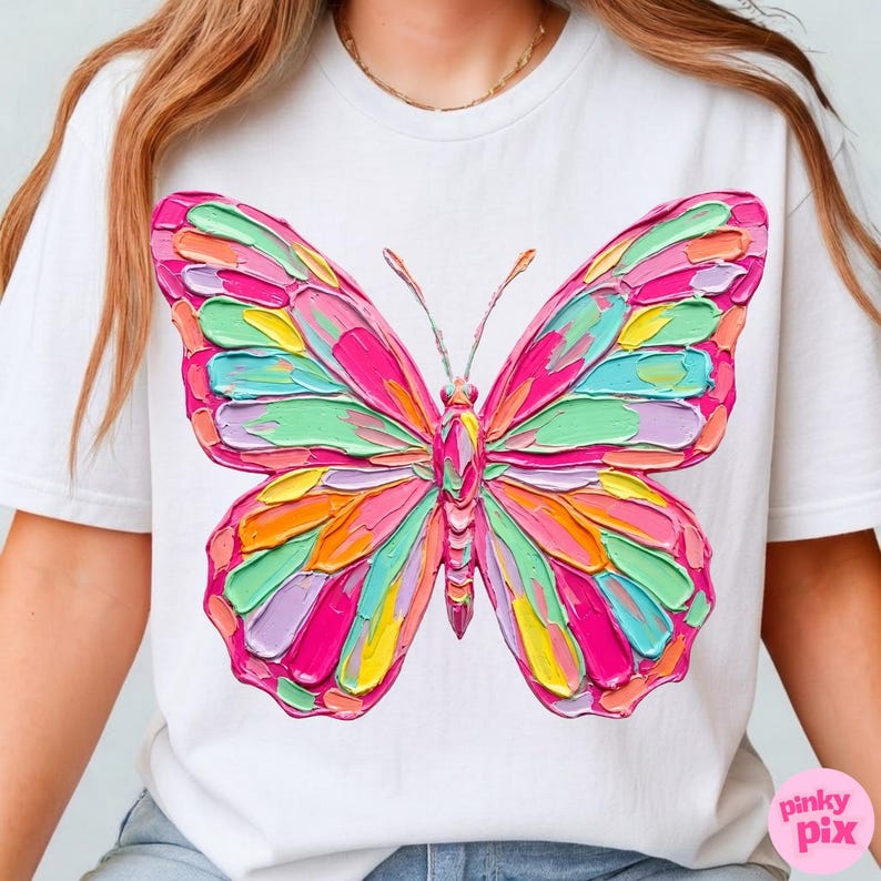 May include: White t-shirt featuring a colorful butterfly design. The butterfly has a textured, painted appearance with shades of pink, green, orange, and yellow. The shirt has a crew neck and short sleeves. The brand name "pinky pix" is in the bottom right corner.