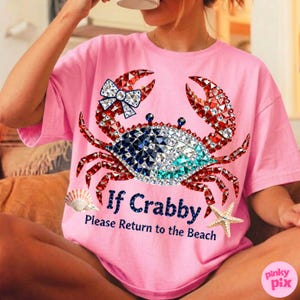 May include: Pink t-shirt featuring a bejeweled crab design with red, blue, and white crystals. The crab has a bow on one claw. Text on the shirt reads "If Crabby, Please Return to the Beach". A seashell and starfish are also on the shirt.