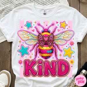May include: White t-shirt featuring a colorful graphic of a bee with a pink bow and the word "KIND" in large pink letters. The bee has yellow and pink stripes, with blue and yellow stars surrounding it. The design is set against a pink striped background.