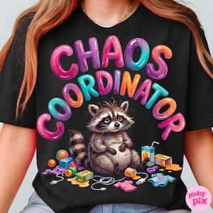 Può includere: T-shirt nera con la scritta "CHAOS COORDINATOR" in lettere colorate. Un procione dei cartoni animati con un'espressione triste è seduto sotto il testo, circondato da giocattoli e scatole di succo. La maglietta ha un girocollo.