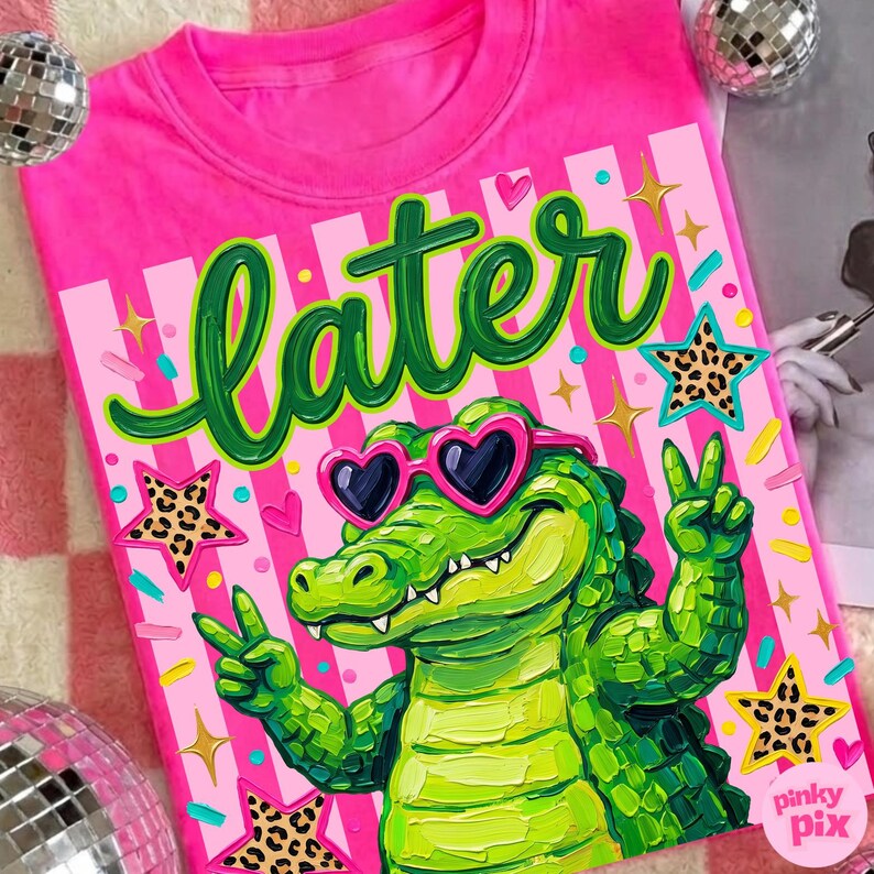 May include: A bright pink t-shirt featuring a green alligator wearing heart-shaped sunglasses and giving a peace sign. The word "later" is written in green script above the alligator. The design includes stars and stripes.