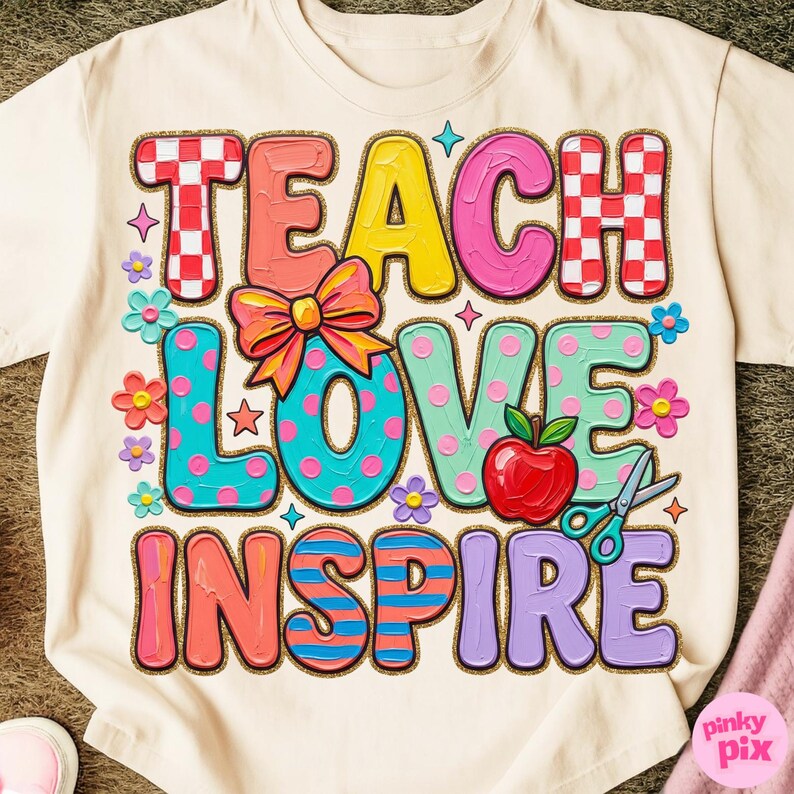 May include: Cream-colored t-shirt with a colorful graphic that reads "TEACH LOVE INSPIRE." The design features a bow, apple, scissors, and flowers. The letters are in various colors and patterns, perfect for teachers.
