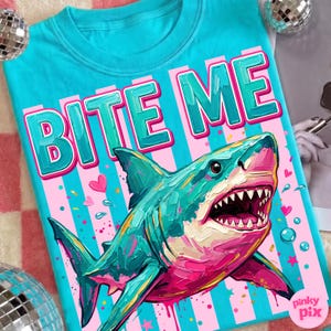 May include: A turquoise t-shirt with the words "BITE ME" in large, pink and green letters. Below the text is a colorful illustration of a shark with its mouth open, set against a pink and blue striped background.