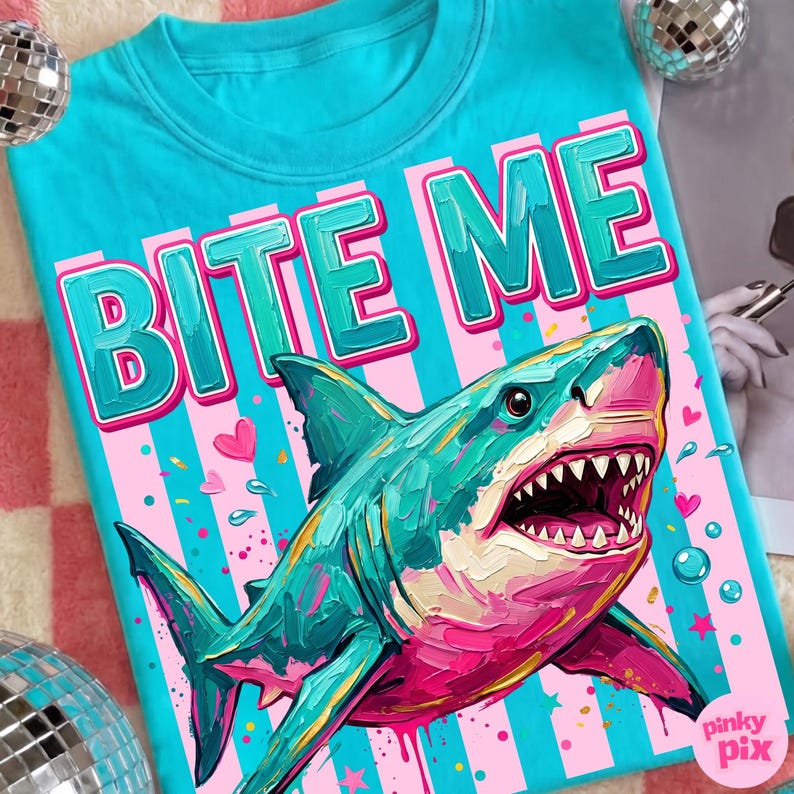 May include: A turquoise t-shirt with the words "BITE ME" in large, pink and green letters. Below the text is a colorful illustration of a shark with its mouth open, set against a pink and blue striped background.